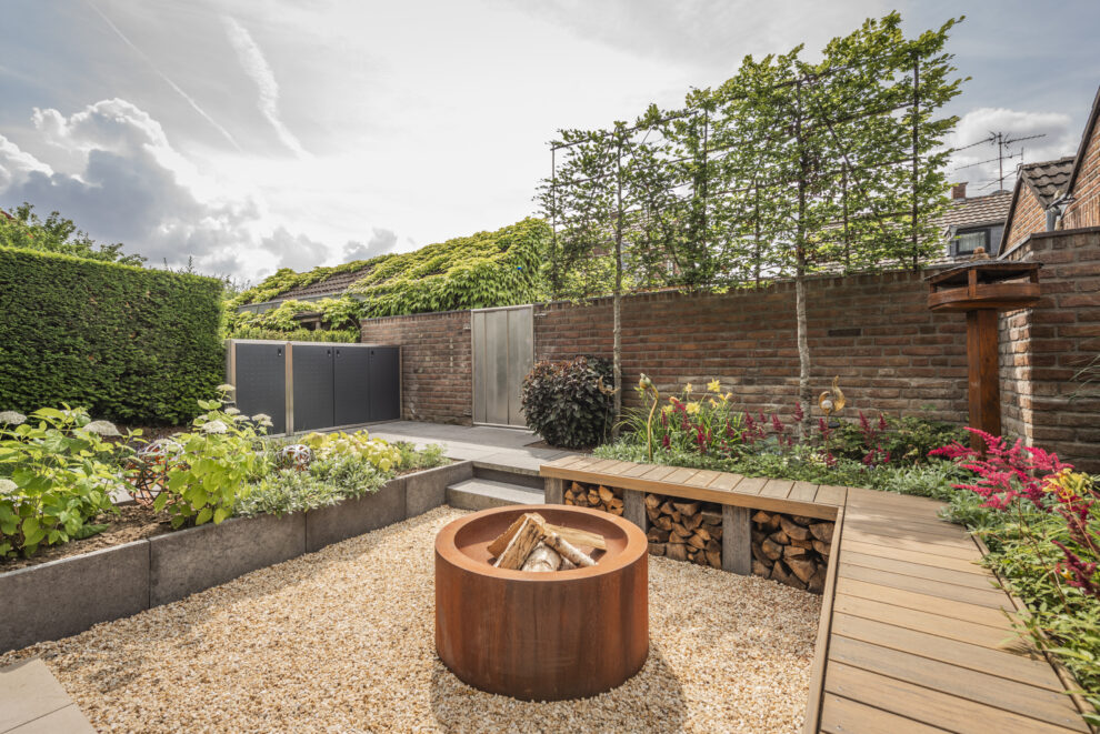 Garten mit Feuerschale aus Cortenstahl, umgeben von einer gepflasterten Terrasse und Pflanzen.