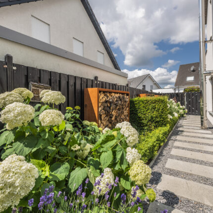 Schmaler Gartenweg mit blühenden Hortensien und Cortenstahl-Holzregal.