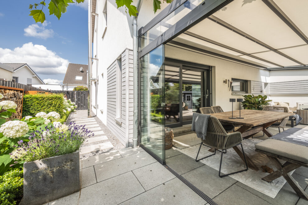 Seitlicher Blick auf die Terrasse mit Essbereich, Glasfront und blühenden Pflanzen.