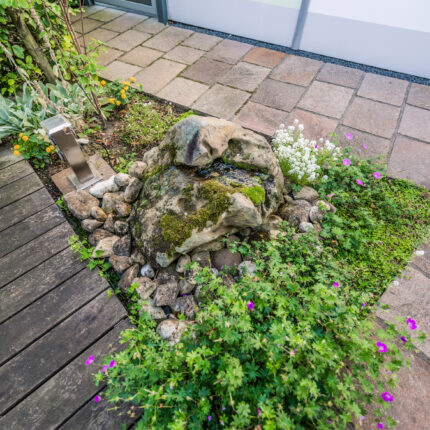 Nahaufnahme eines kleinen Gartenteichs mit sprudelndem Wasser über moosbedeckten Steinen, der dem Garten ein natürliches Wasserspiel verleiht.